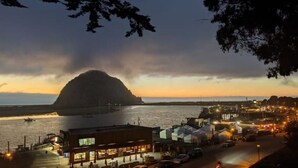 Un pueblo costero al anochecer, con una colina destacada en el fondo, un puerto deportivo con barcos y una zona ribereña iluminada con edificios y tiendas.