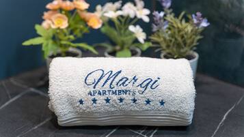 Une serviette pliée ornée d’un texte brodé « Margo Apartments » et une rangée de plantes en pot, avec diverses fleurs en arrière-plan.