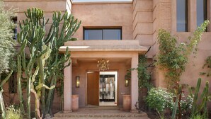 Un edificio con un cactus grande al frente, un acceso con puerta de vidrio y una ventana pequeña en el piso superior.