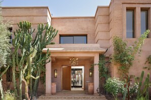 Un edificio con un cactus grande al frente, un acceso con puerta de vidrio y una ventana pequeña en el piso superior.