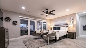 A bedroom with a bed, two armchairs, a nightstand, a ceiling fan, and a large window with a view. - Stunning Lake View, Heated Pool, Single Story! (Boulder City)