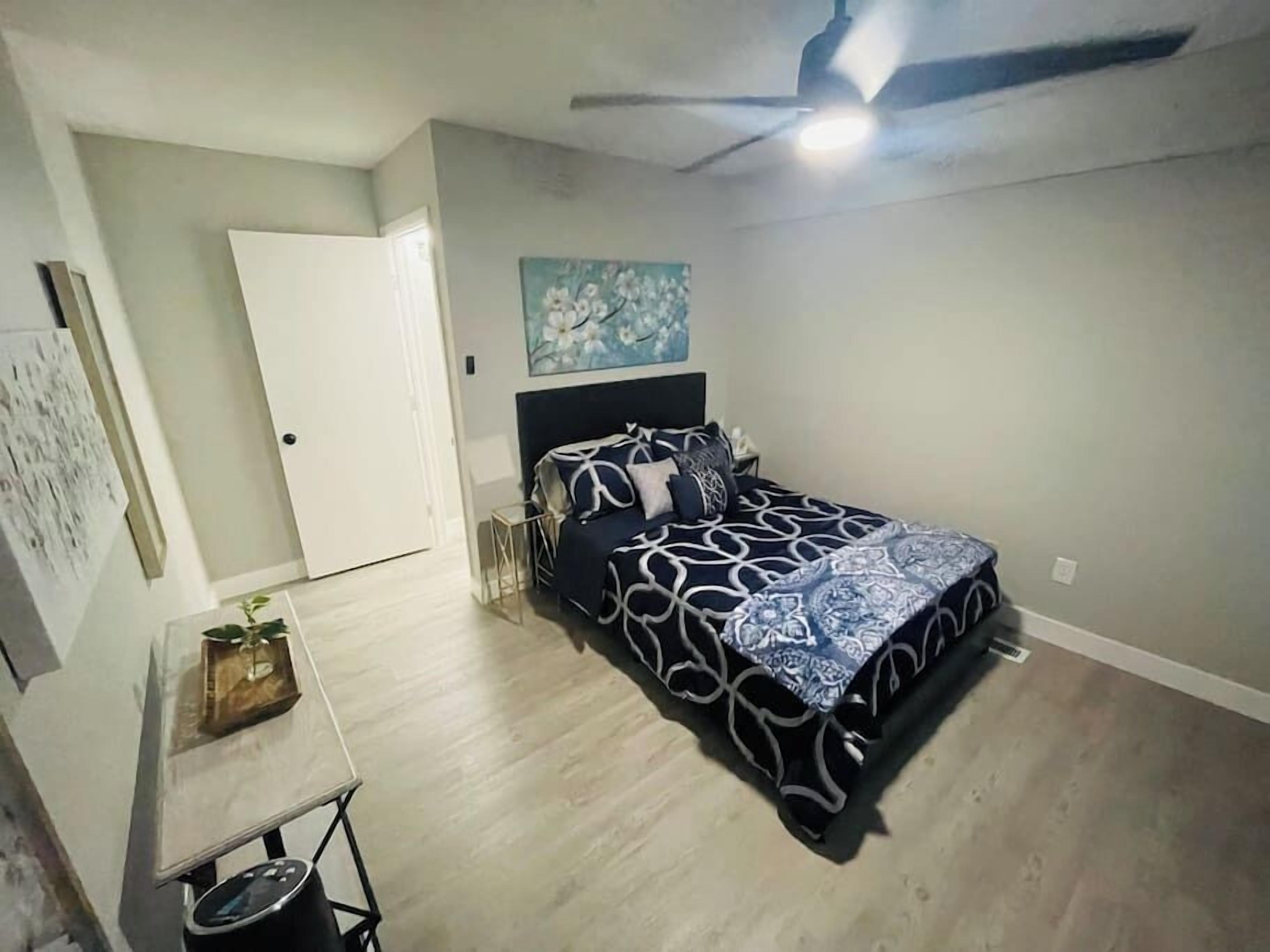 Un dormitorio con cama, ventilador de techo, un cuadro en la pared y una mesita con una planta.
