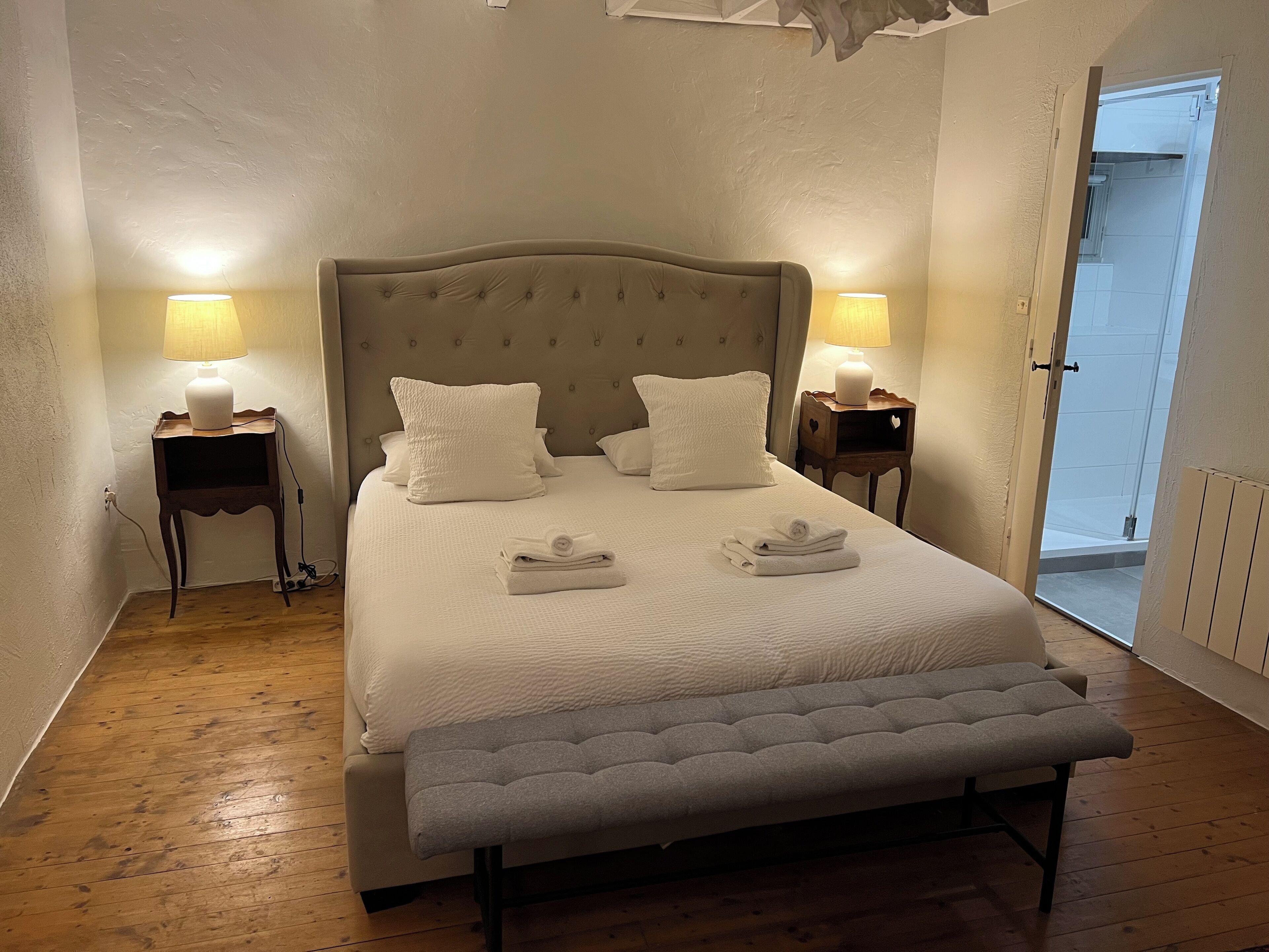 Un lit bien fait, avec du linge de lit blanc et des oreillers, deux tables de chevet avec des lampes, un banc au pied du lit et une vue sur la salle de bain par une porte ouverte.