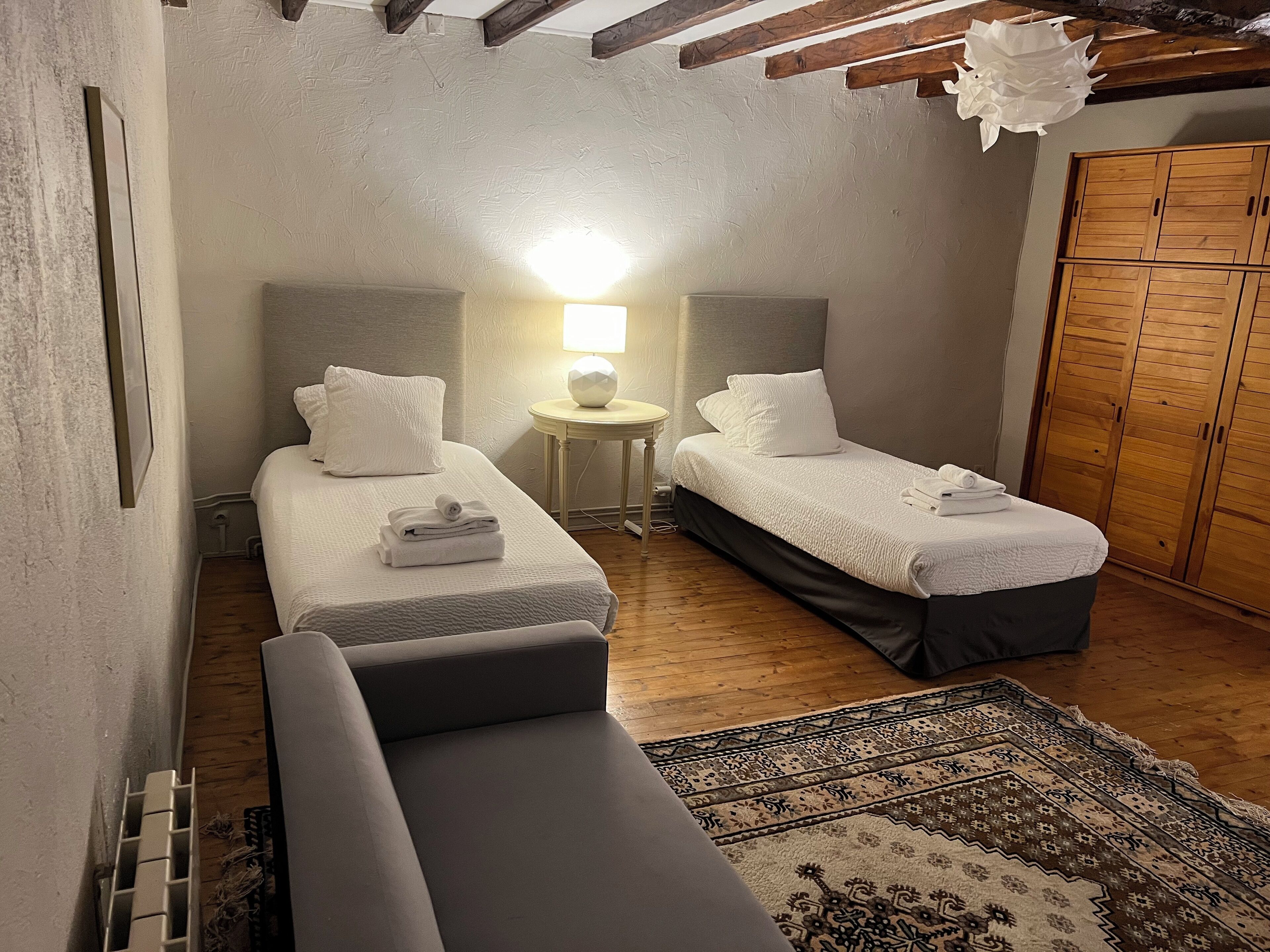 Um quarto pequeno e acolhedor com duas camas de solteiro, uma mesinha de cabeceira com abajur, um guarda-roupa de madeira e um tapete estampado.