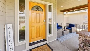 Una entrada de casa con puerta de madera, felpudo y porche con mobiliario exterior.