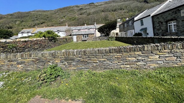 A stone wall with a grassy foreground and buildings in the background. - The Brewhouse Boscastle Harbour (Boscastle)