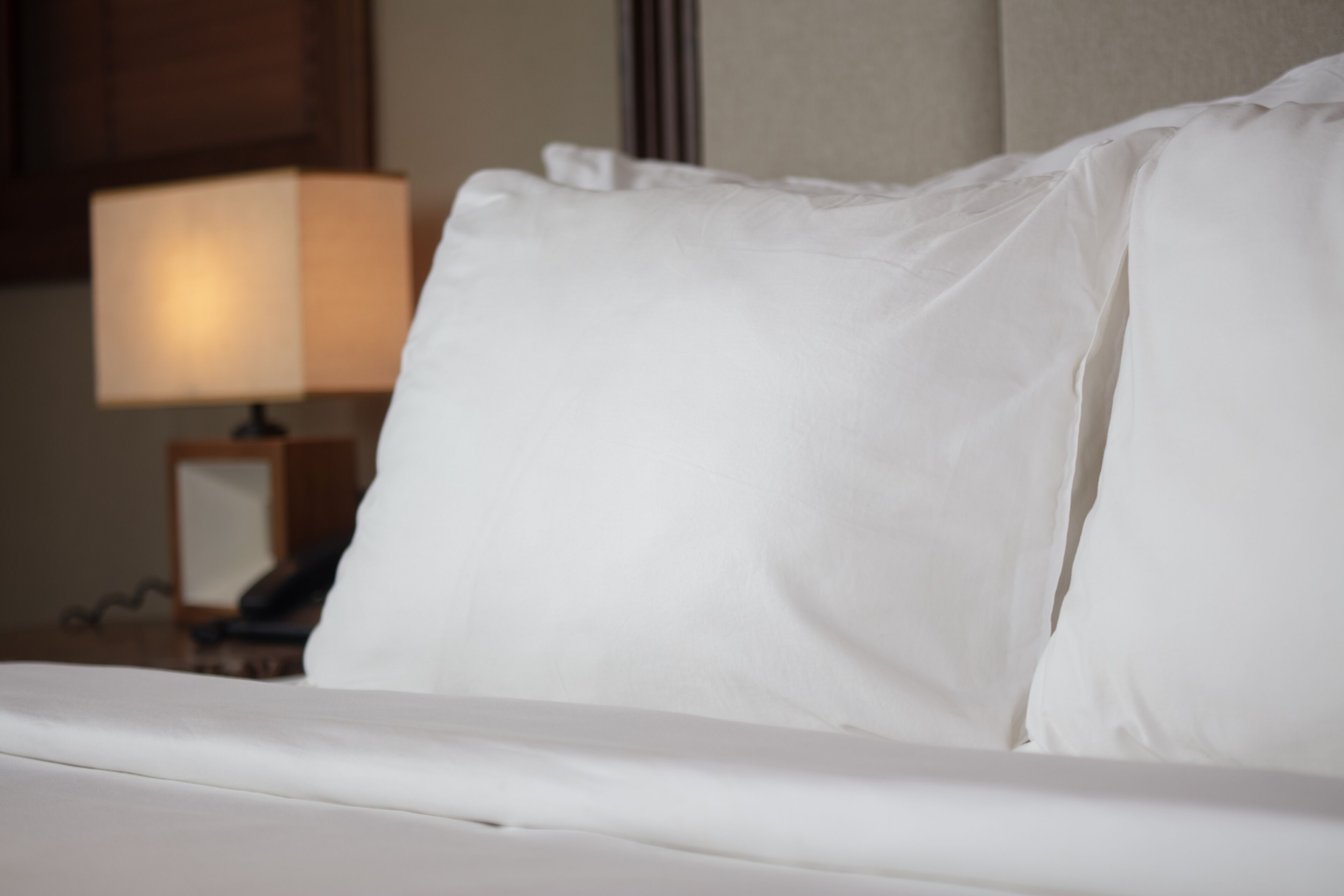 A neatly made bed with white linens and pillows, a bedside lamp, and a wooden nightstand with a framed picture.