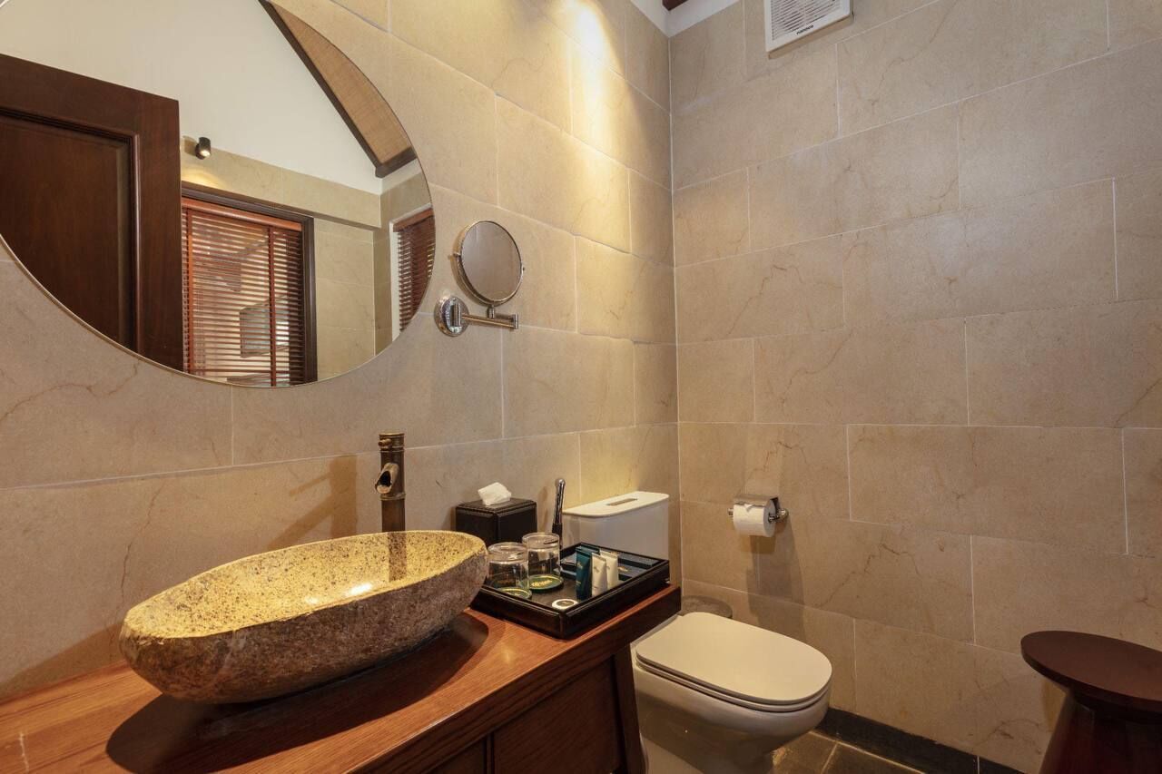 A bathroom with a stone sink, a round mirror, a toilet, and a wooden vanity.
