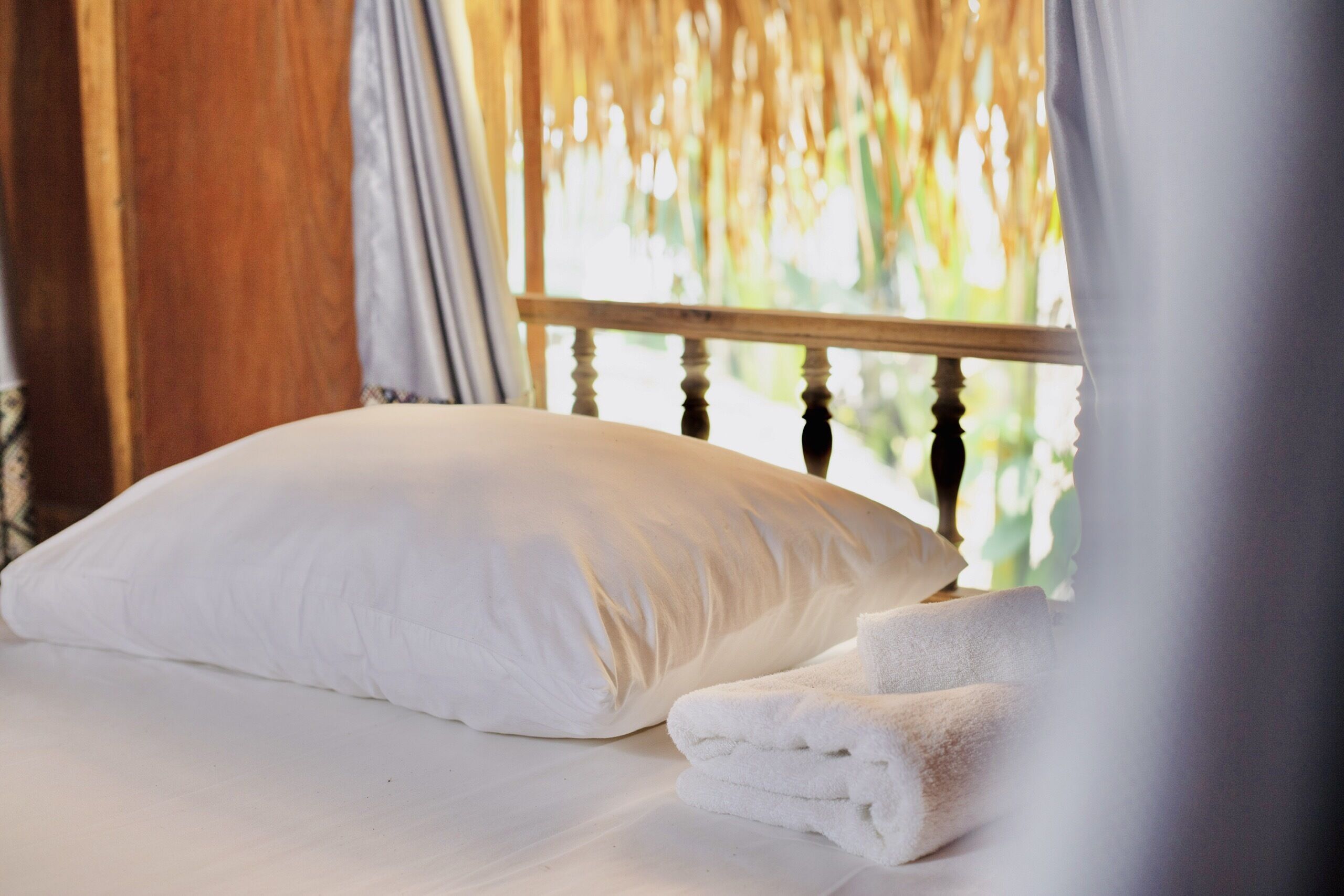 Una cama con almohada blanca y toallas dobladas, cabecera de madera y vista a la vegetación a través de una ventana con cortinas.
