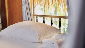 Una cama con almohada blanca y toallas dobladas, cabecera de madera y vista a la vegetación a través de una ventana con cortinas.