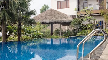 Une piscine dotée d’une structure de toit en chaume, entourée d’une végétation luxuriante et avec un immeuble à plusieurs étages en arrière-plan.