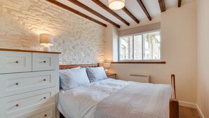 A bedroom with a stone wall, a bed with white bedding, wooden beams, a nightstand with a lamp, and a window with a view of buildings. - Grade II Old Engine House, on the SW Coast Path (Noss Mayo)