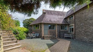 A stone building with a brick patio, a bench, and a staircase leading up to the entrance. - Grade II Old Engine House, on the SW Coast Path (Noss Mayo)