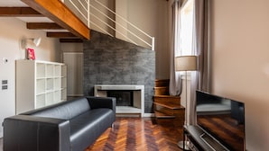 Uma sala de estar moderna com um sofá de couro preto, uma televisão de tela plana, uma lareira, piso de madeira e uma escada com vigas aparentes.