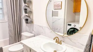 A bathroom with a white marble sink, gold faucets, a round mirror with a gold frame, a toilet, and a shower area with grey curtains. - Lovely Home! Great Location! The Nature House (Springfield)