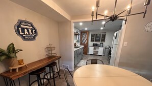 A dining area with a wooden table and bar stools, a wall-mounted sign reading "Welcome to Lake House," and a kitchen visible through an open doorway.