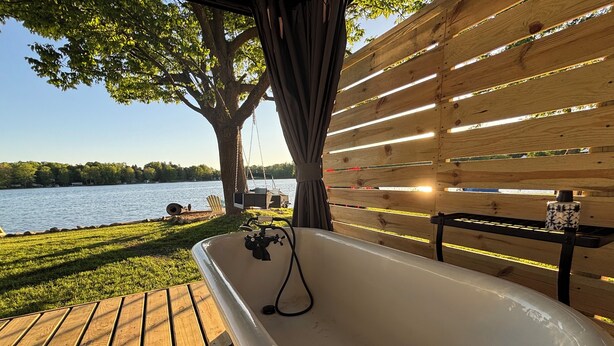 A wooden deck with a bathtub, a table with a mug, and a view of a lake.