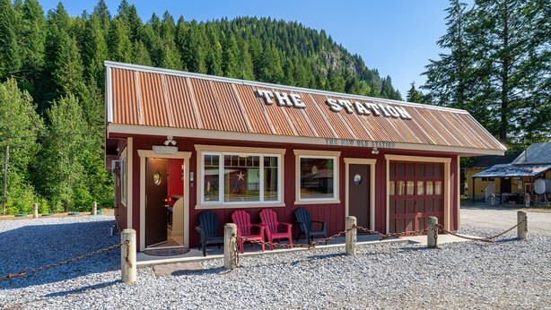 Ein rustikales Gebäude mit dem Schriftzug "The Station", umgeben von Kies und mit Holzstühlen vor der Fassade.