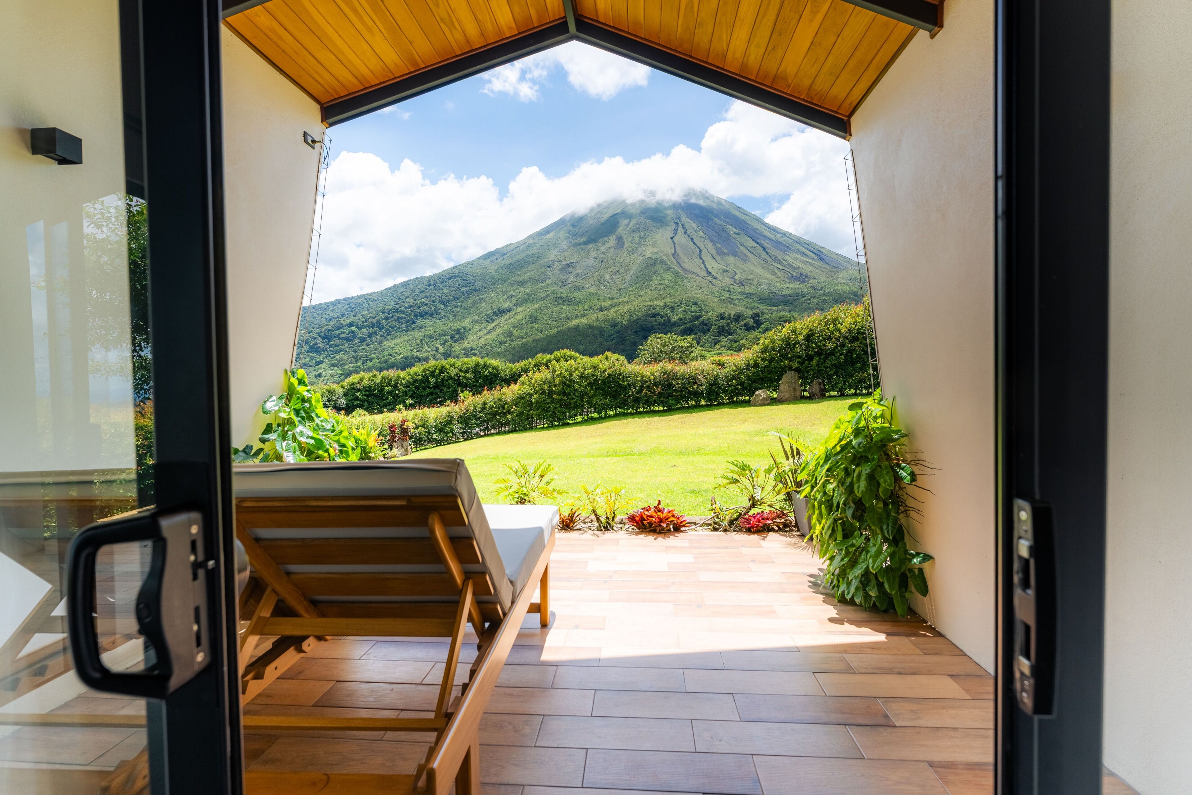 A room with a wooden ceiling, a view of a mountain, a garden with plants, and a wooden chair.