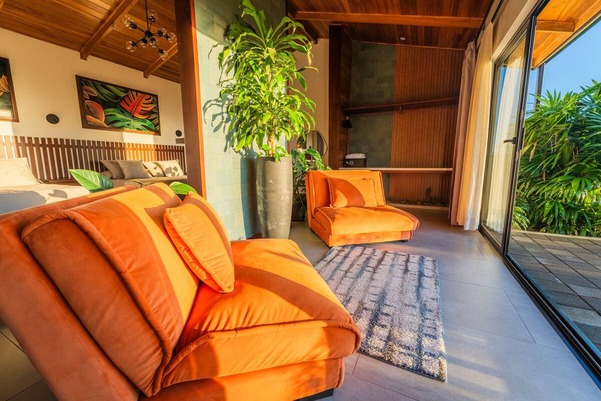 A modern living room with two orange sofas, a large potted plant, and a view of the outdoors.