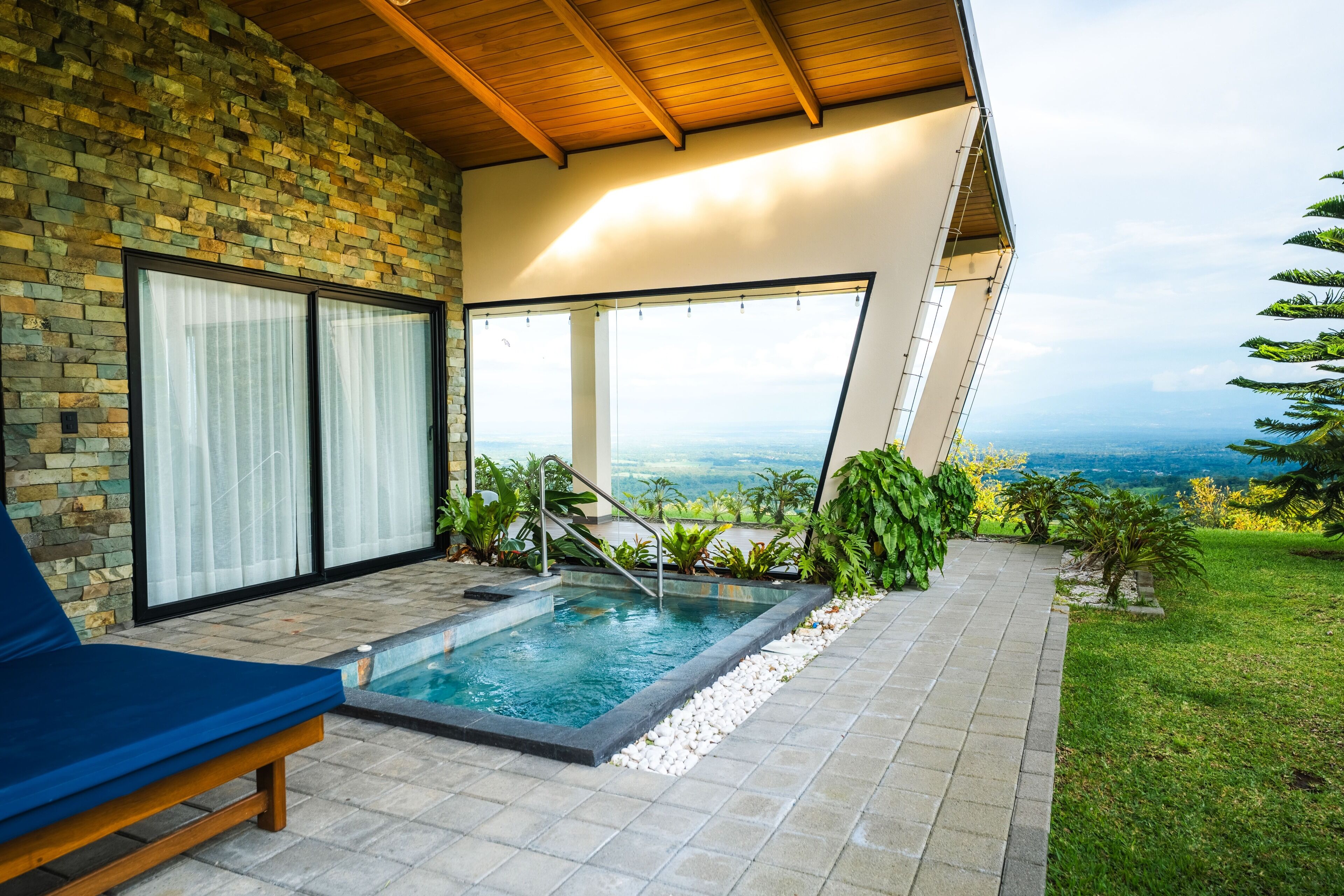 A modern outdoor pool area with a stone wall, a blue lounge chair, and a view of the landscape.