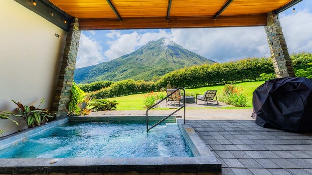 A pool area with a view of a mountain, stone pillars, and outdoor furniture. - Villas Jawara (La Fortuna)