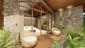 A modern outdoor seating area with wicker chairs, a wooden deck, and stone walls. - Villas Jawara (La Fortuna)