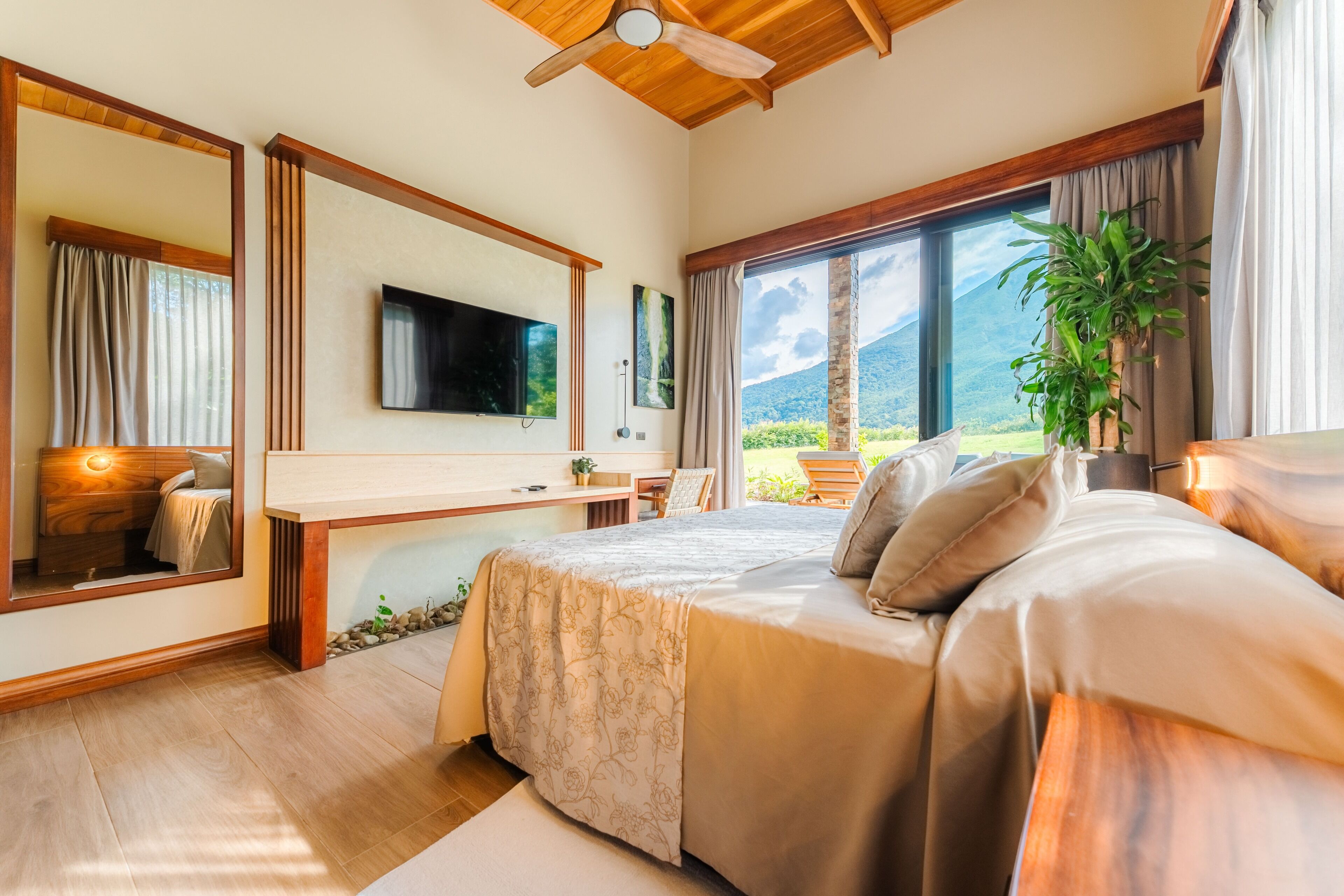A bedroom with a large bed, a desk, a TV, and a view of the outdoors.