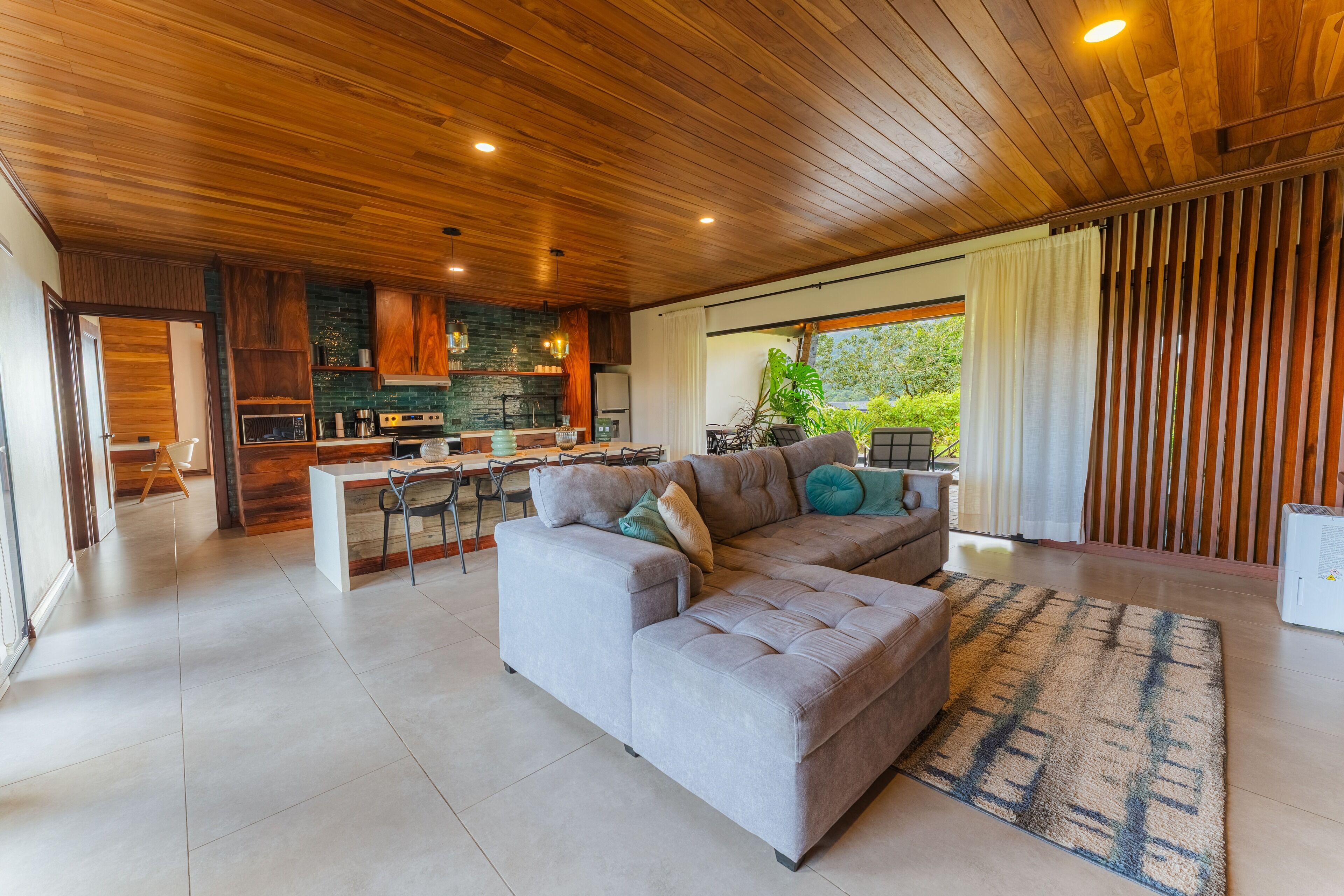 A modern living room with a grey sectional sofa, a kitchen with wooden cabinets, and a large window with curtains.