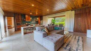 A modern living room with a grey sectional sofa, a kitchen with wooden cabinets, and a large window with curtains.