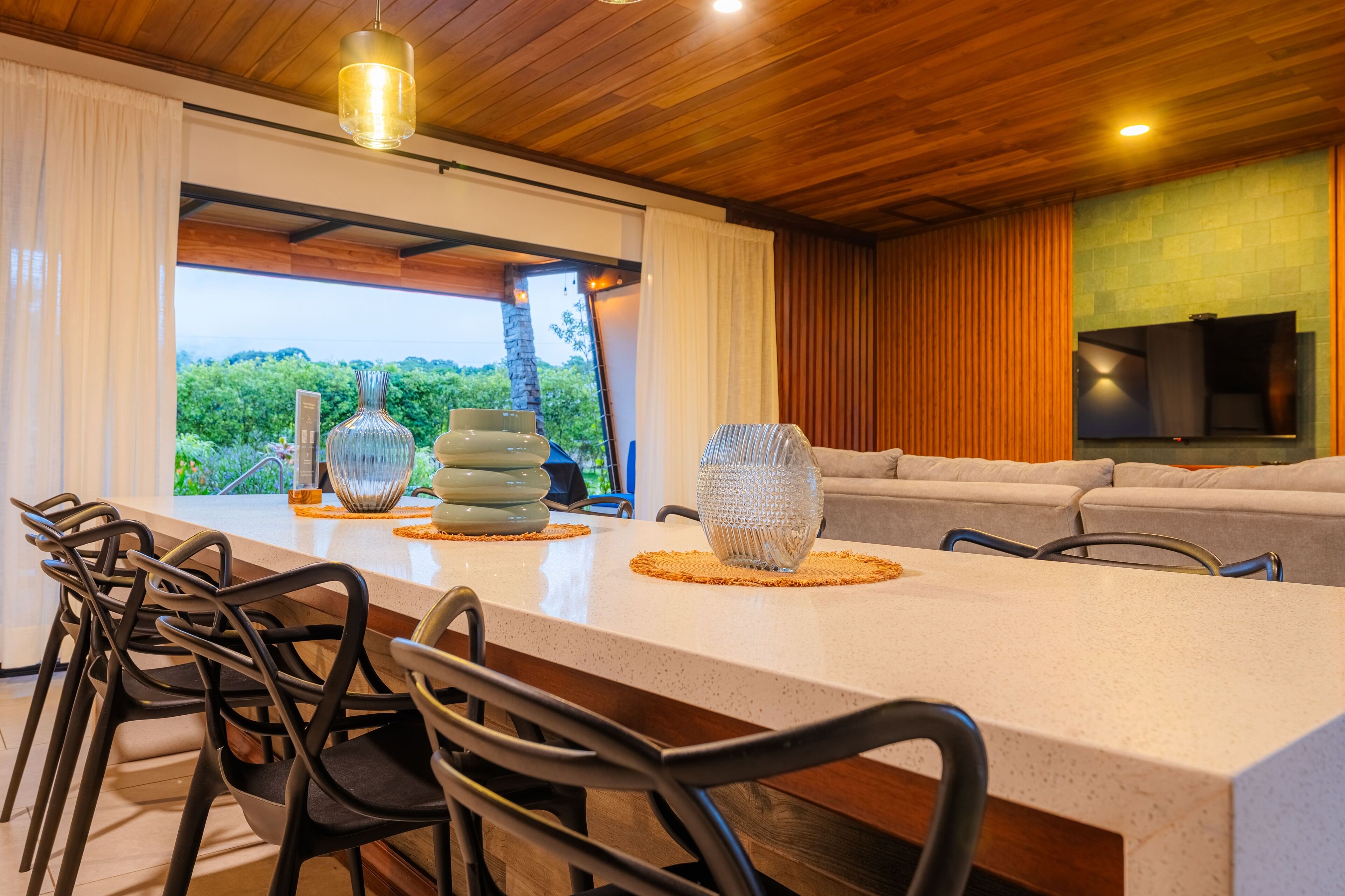 A modern kitchen with a large island, black chairs, and a view of greenery outside.