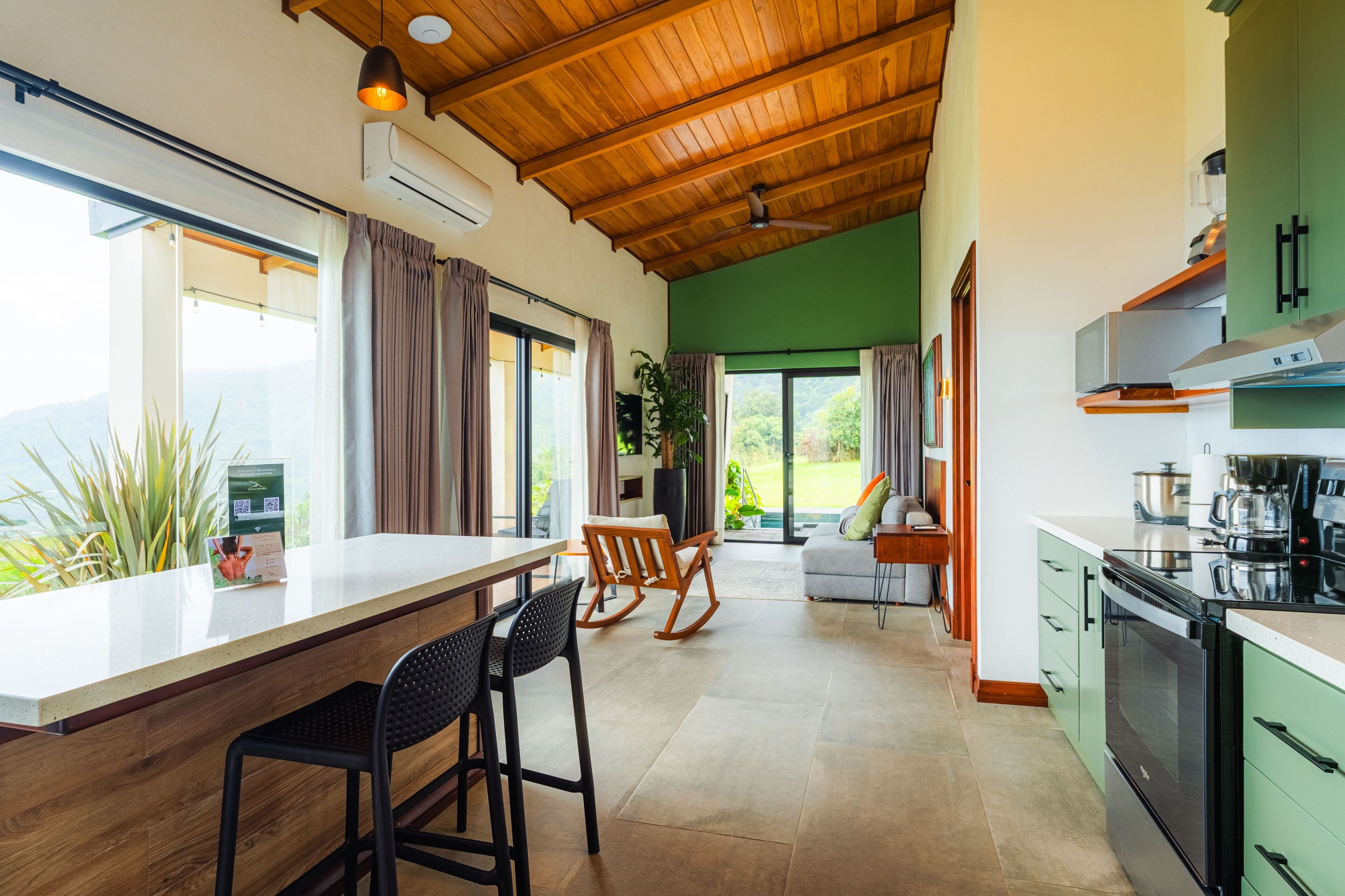A modern kitchen with a wooden ceiling, green cabinets, and a large window overlooking a garden.