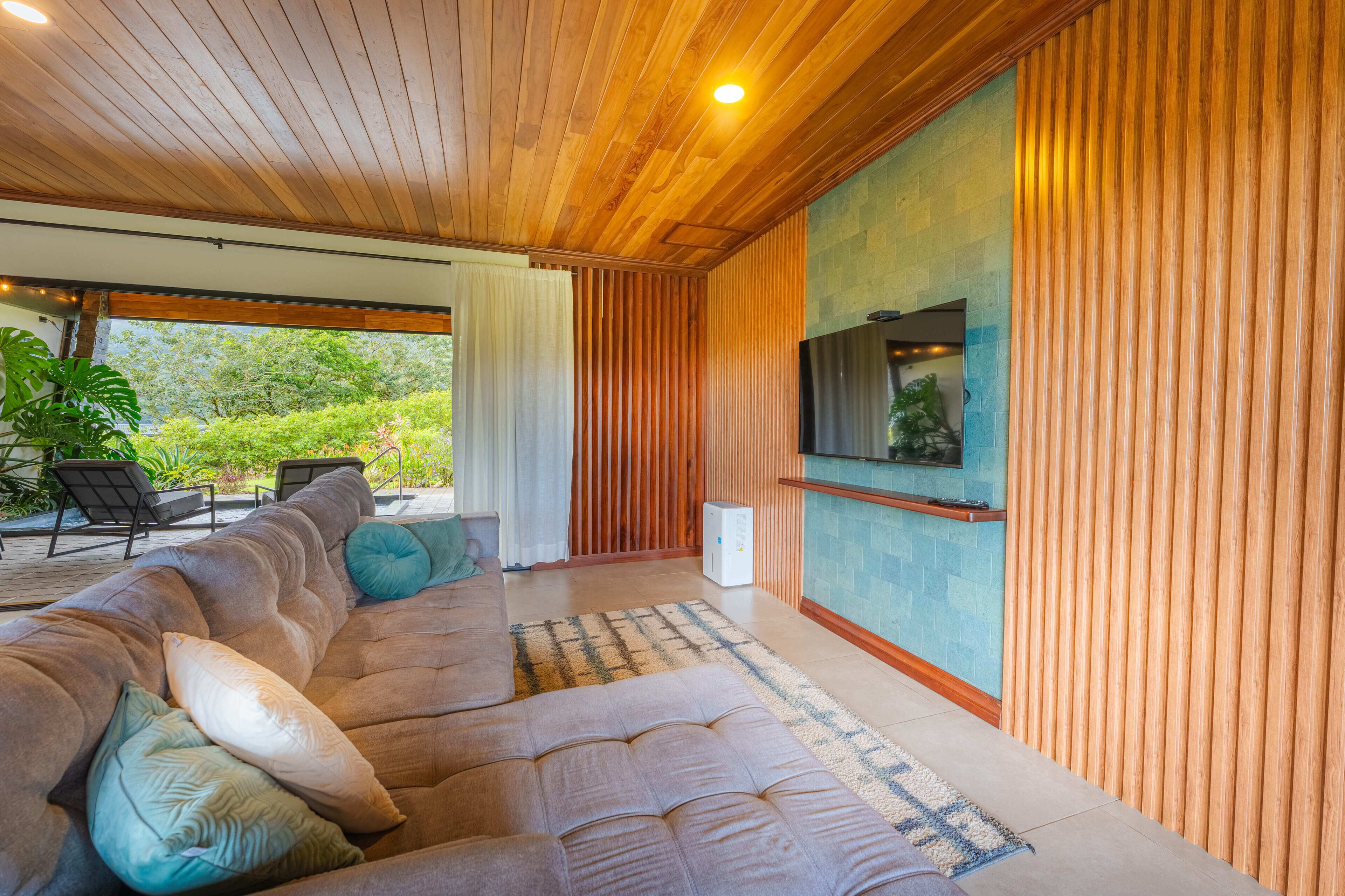 A modern living room with a large sectional sofa, a flat-screen TV mounted on a wooden wall, and a view of greenery outside.