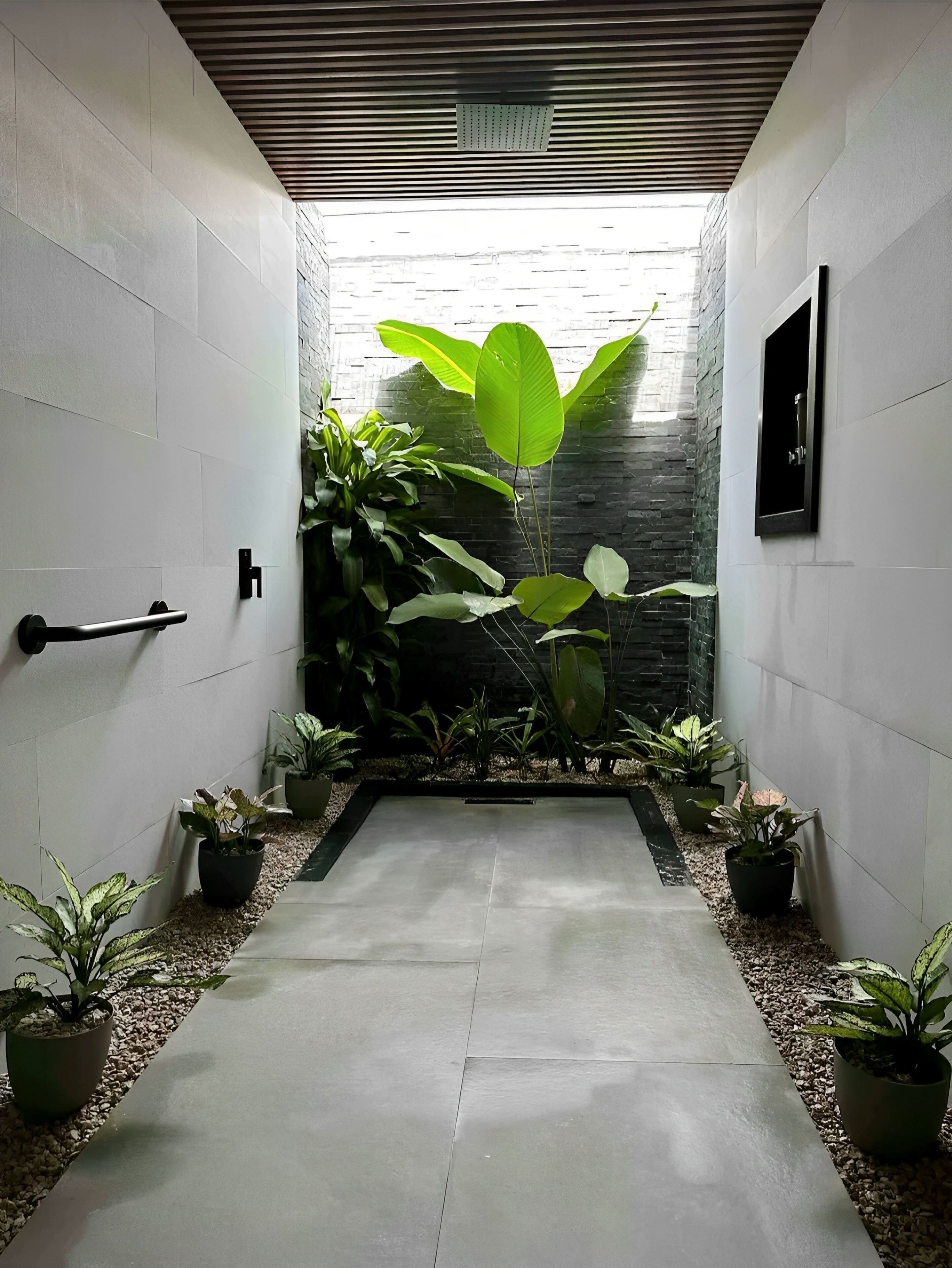 A modern hallway with a concrete floor, white walls, and a stone wall at the end. There are potted plants on either side and a black door handle on the left.
