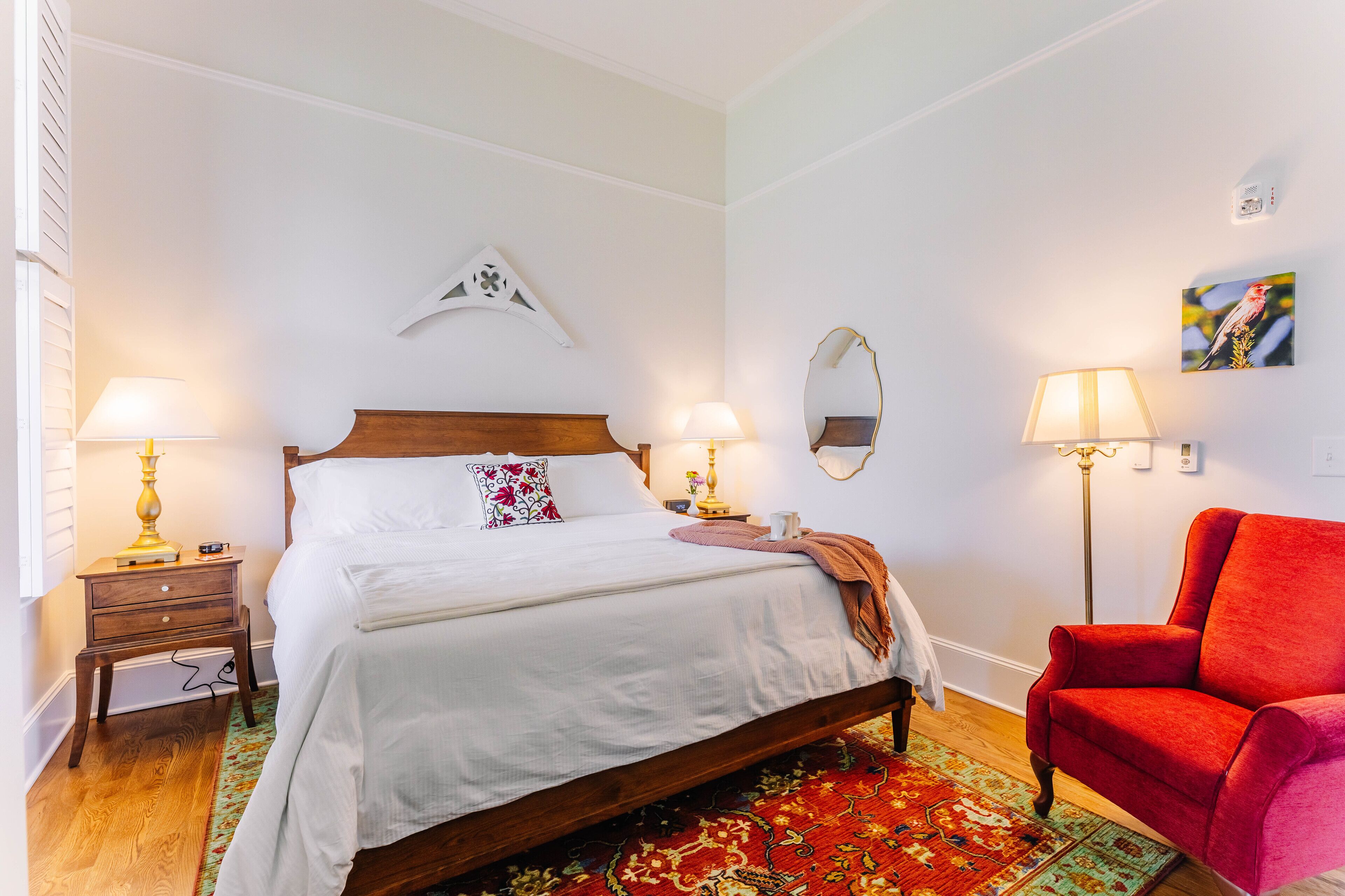 A neatly arranged bedroom with a wooden bed, two bedside tables with lamps, a red chair, a mirror, and a wall-mounted artwork.
