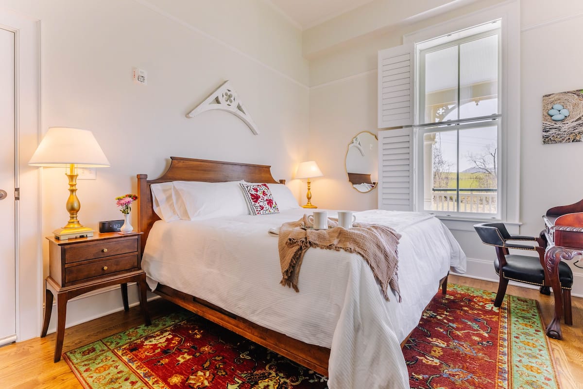 A neatly made bed with a white comforter and a decorative throw blanket. There are two bedside tables with lamps and a small vase with flowers. A colorful rug covers the floor, and a wooden chair is positioned near the window.