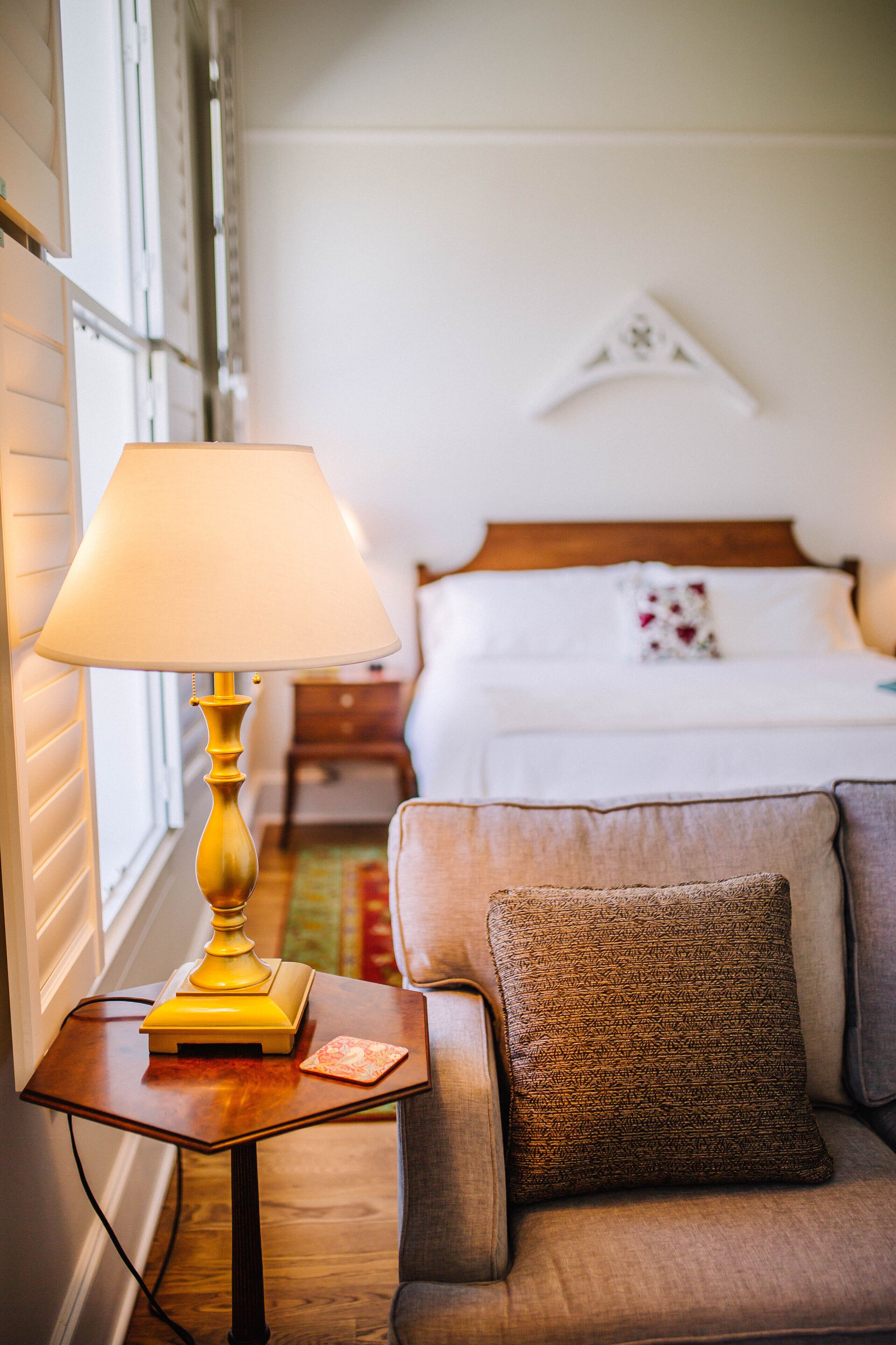 A hotel room with a bed, a bedside table with a lamp, and a sofa with a cushion.