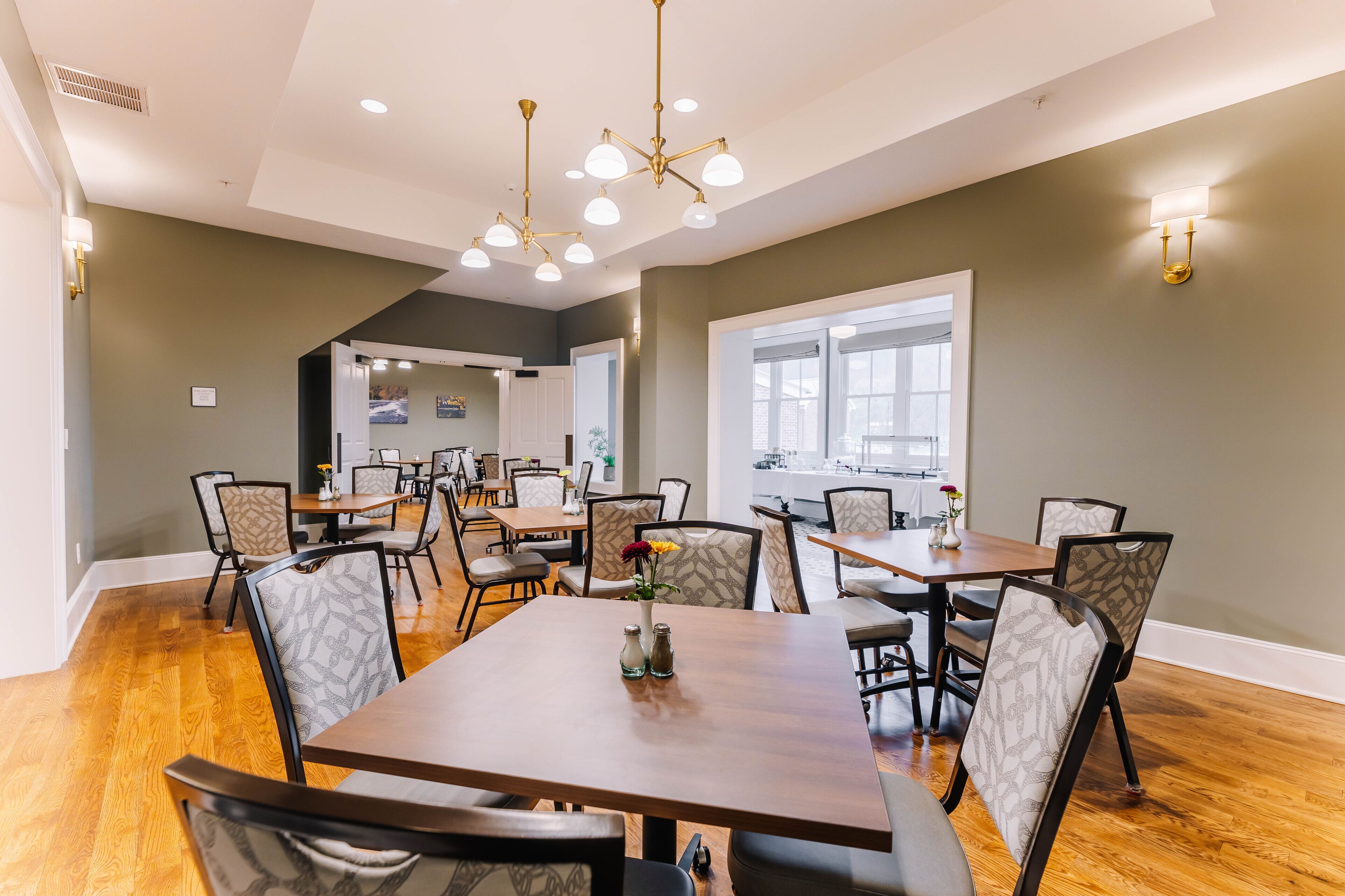 A spacious dining area with multiple tables and chairs, a modern chandelier, and large windows allowing natural light.