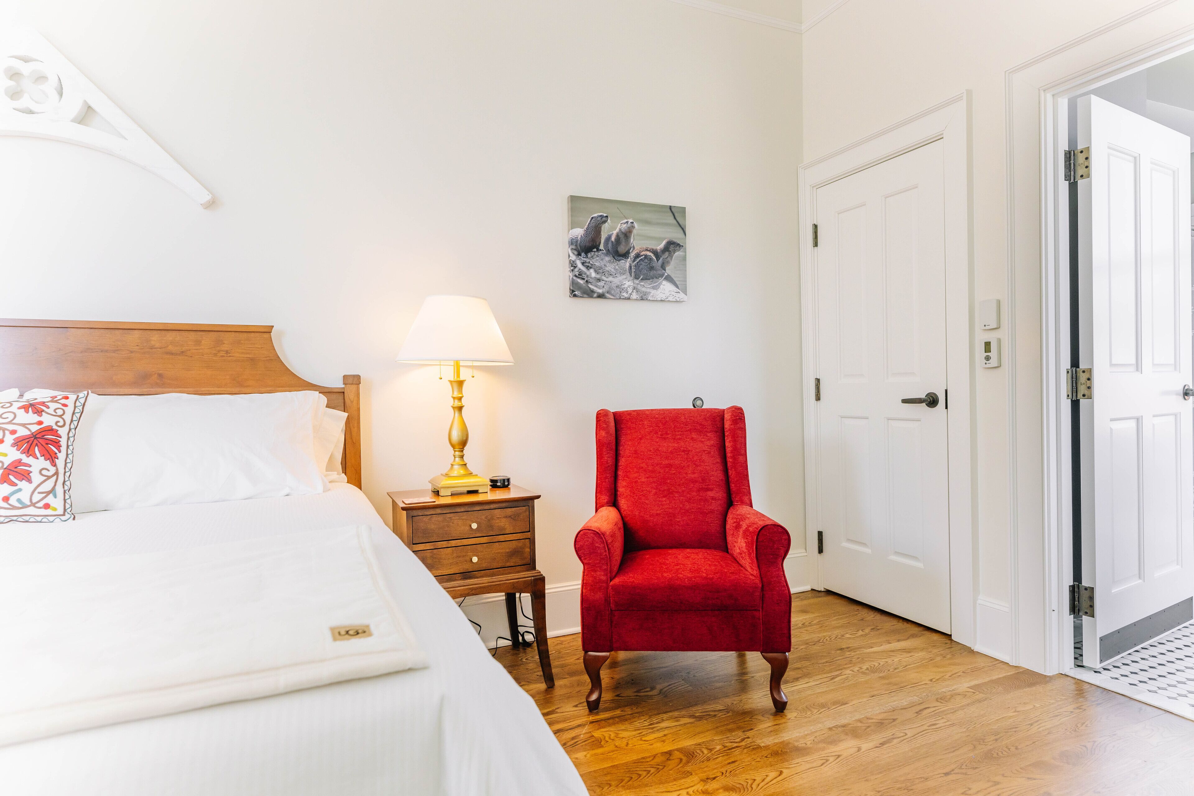 A hotel room with a bed, a red armchair, a bedside table with a lamp, a picture on the wall, and a door.