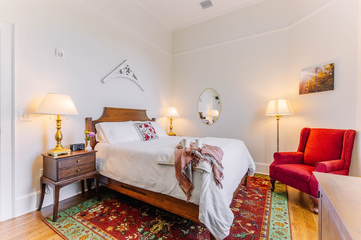 A neatly arranged bedroom with a bed, bedside tables, lamps, a red armchair, and a patterned rug.