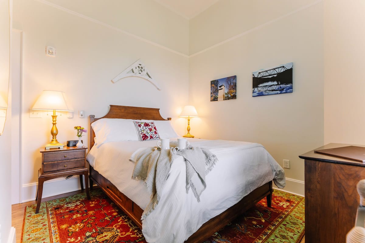 A neatly made bed with a white comforter and two pillows, flanked by two bedside tables with lamps and books. A small decorative item is on the left table, and a wall-mounted shelf is above the bed.