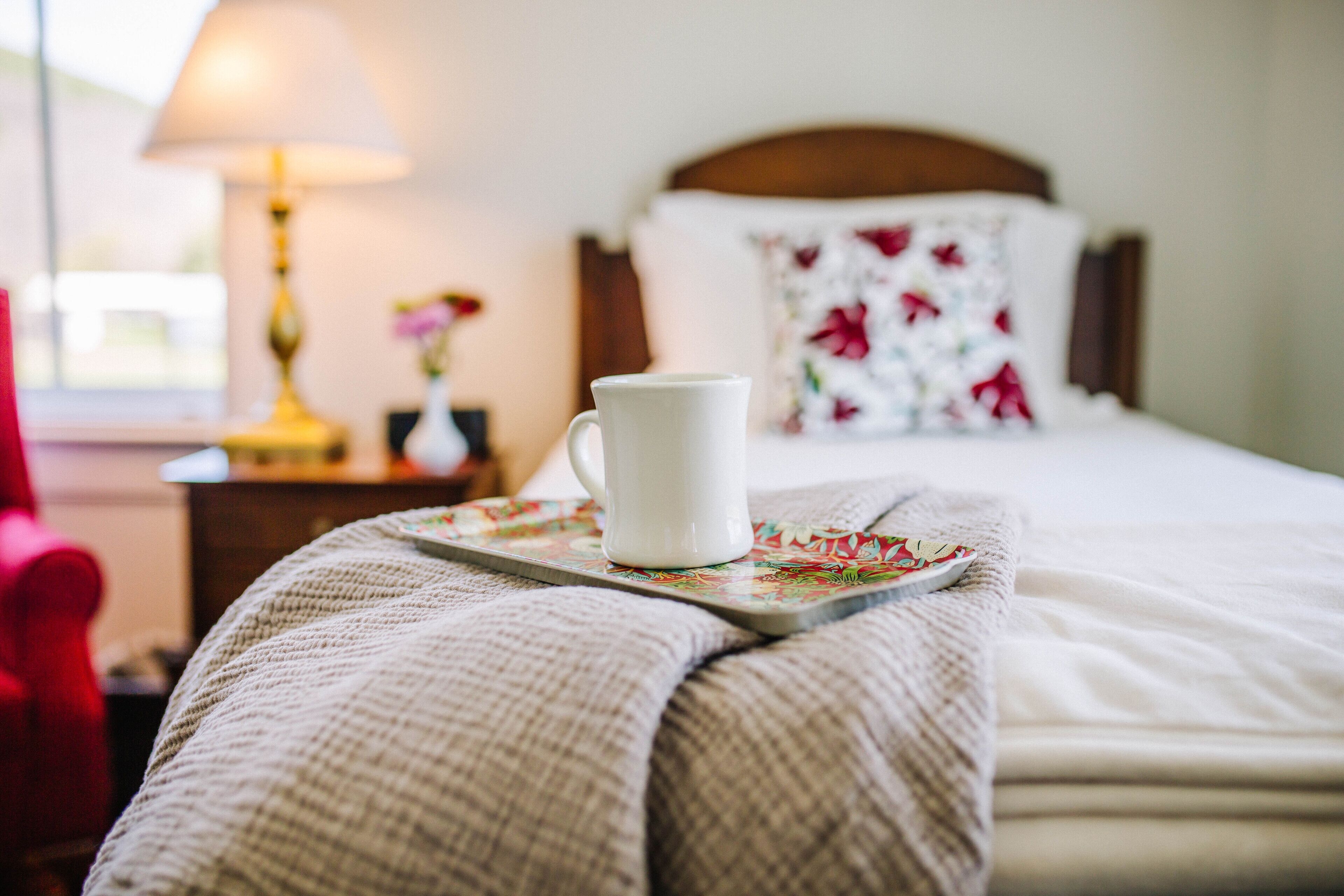 A neatly made bed with a white pillow and a floral-patterned pillow, a white mug on a coaster, and a lamp on a bedside table.