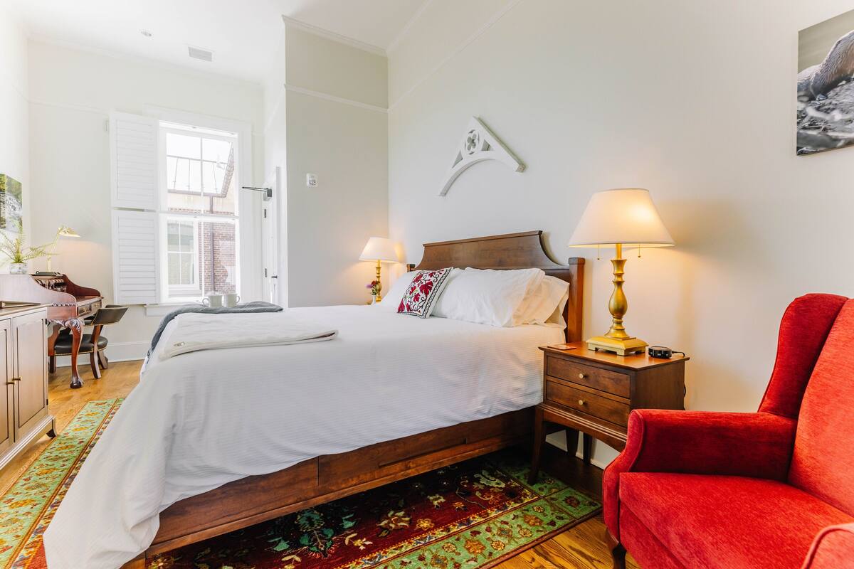 A bedroom with a bed, two bedside tables with lamps, a red chair, a desk with a chair, and a window with shutters.