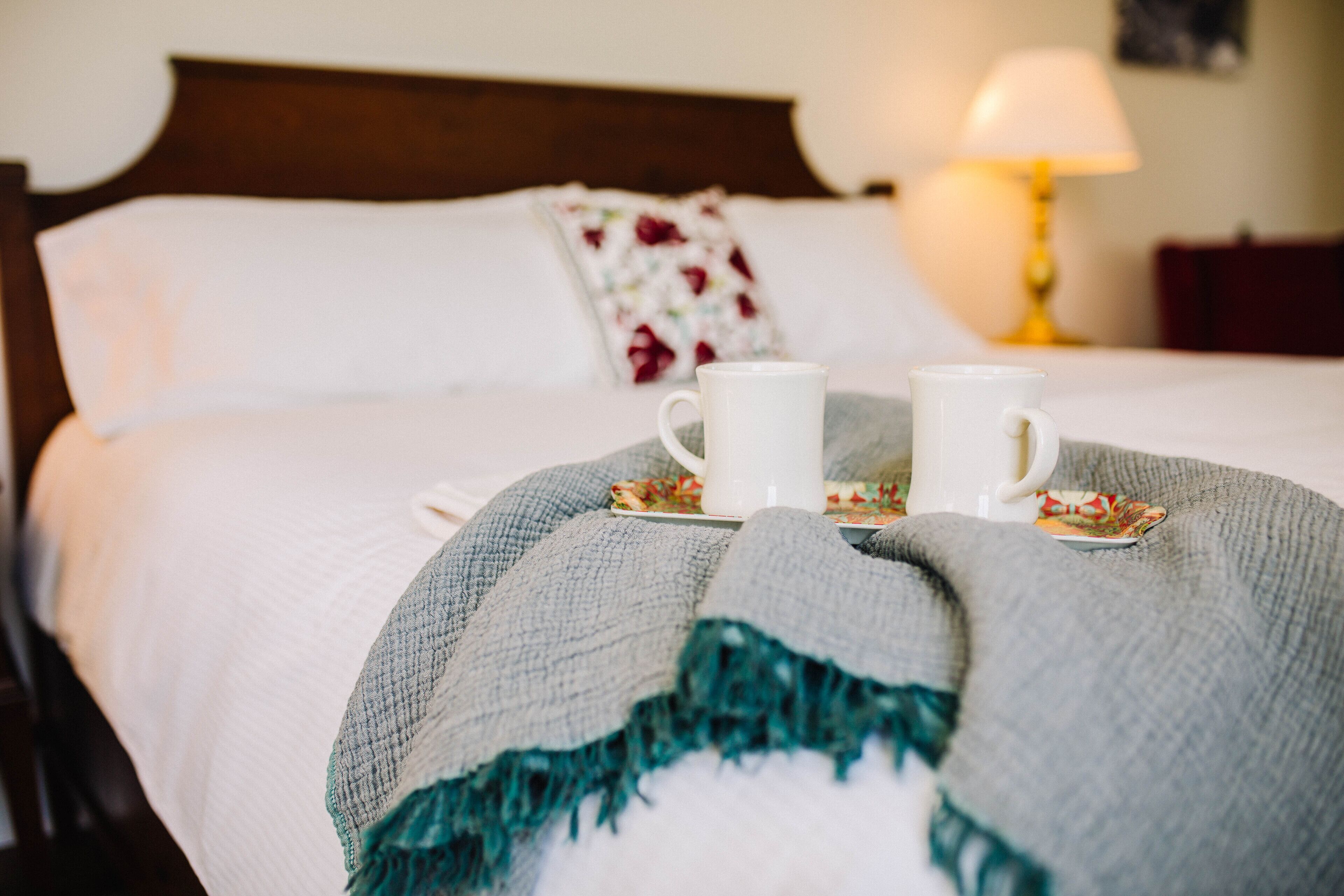 Una cama bien tendida con un cojín floral, dos tazas blancas en una bandeja y una lámpara de noche.