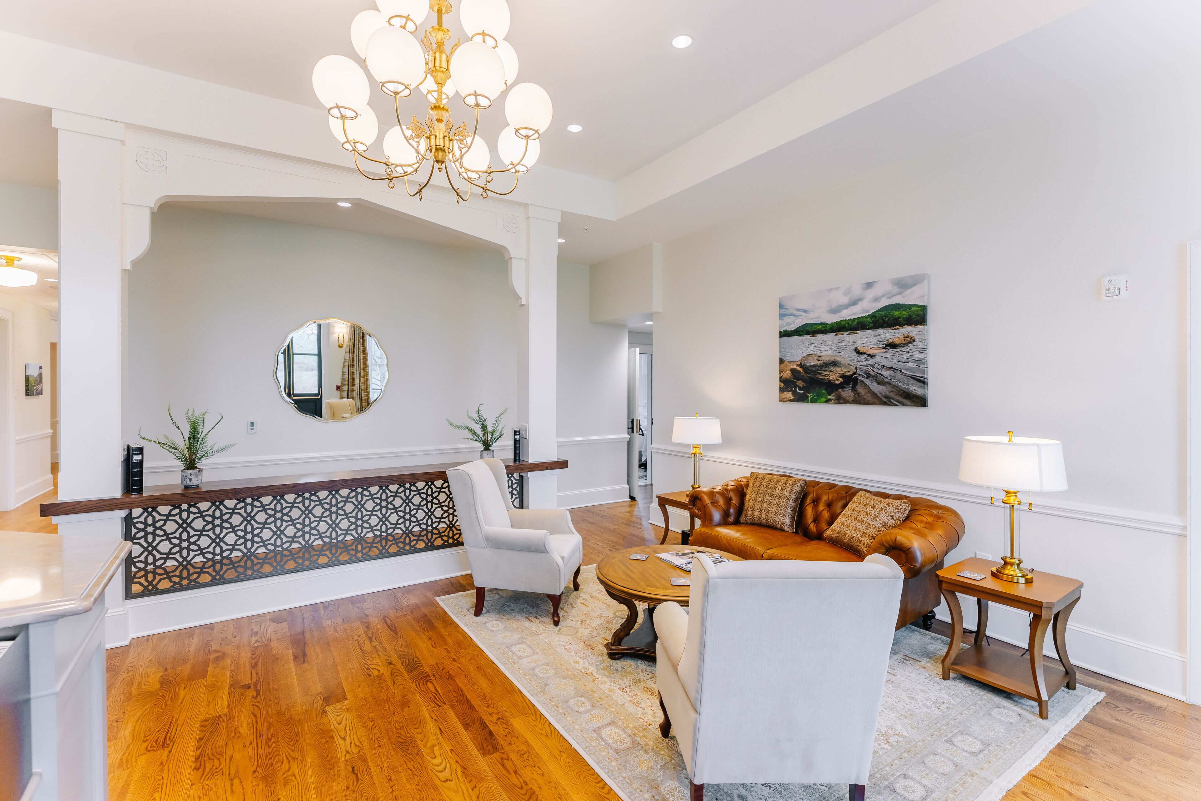 A spacious living room with a wooden floor, a brown leather sofa, a white armchair, a round coffee table, and a chandelier.