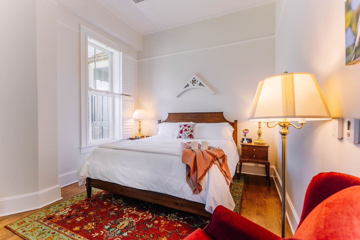 A bedroom with a bed, two bedside tables, a nightstand, a red chair, a floor lamp, and a window with white shutters.