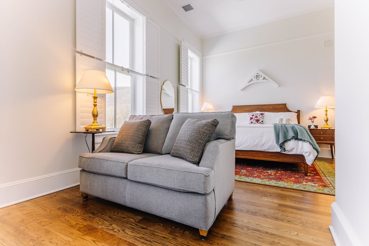 A bedroom with a grey sofa, a wooden floor, a bed with a white headboard, and a window with shutters.