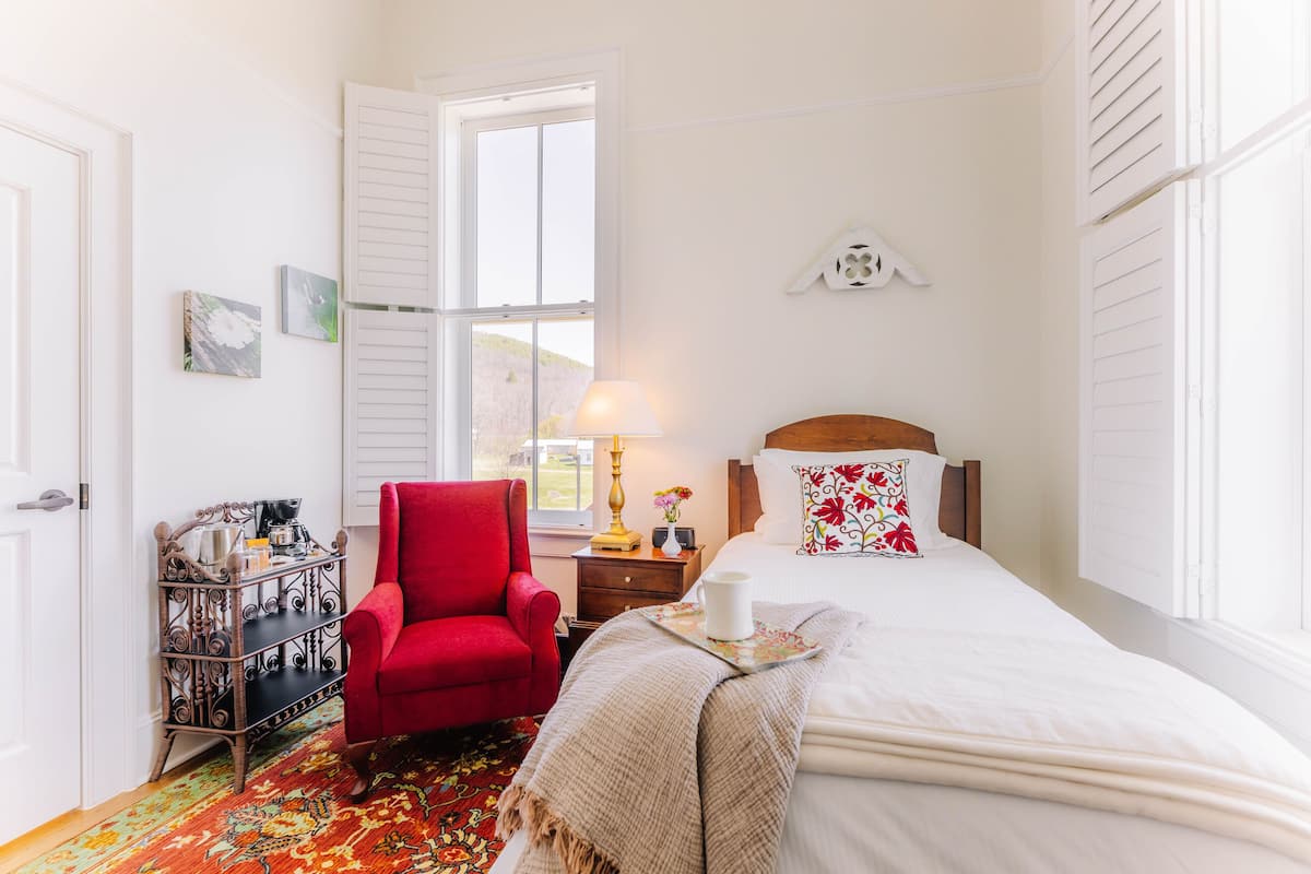 A bedroom with a large bed, a red armchair, a wooden side table, and a window with shutters.