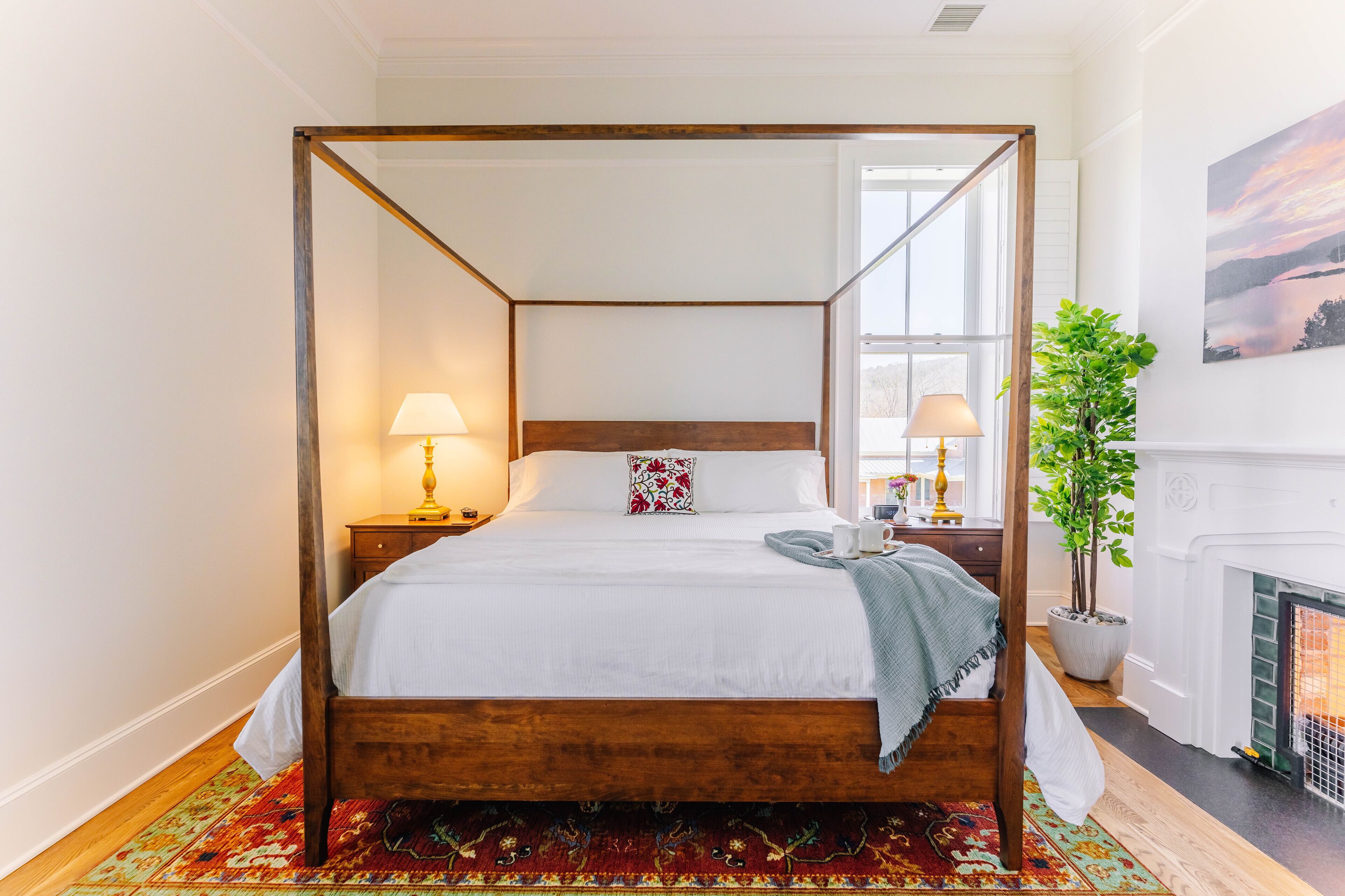 A four-poster bed with a wooden frame, white bedding, and a patterned throw. There is a nightstand with a lamp, a potted plant, and a painting on the wall.