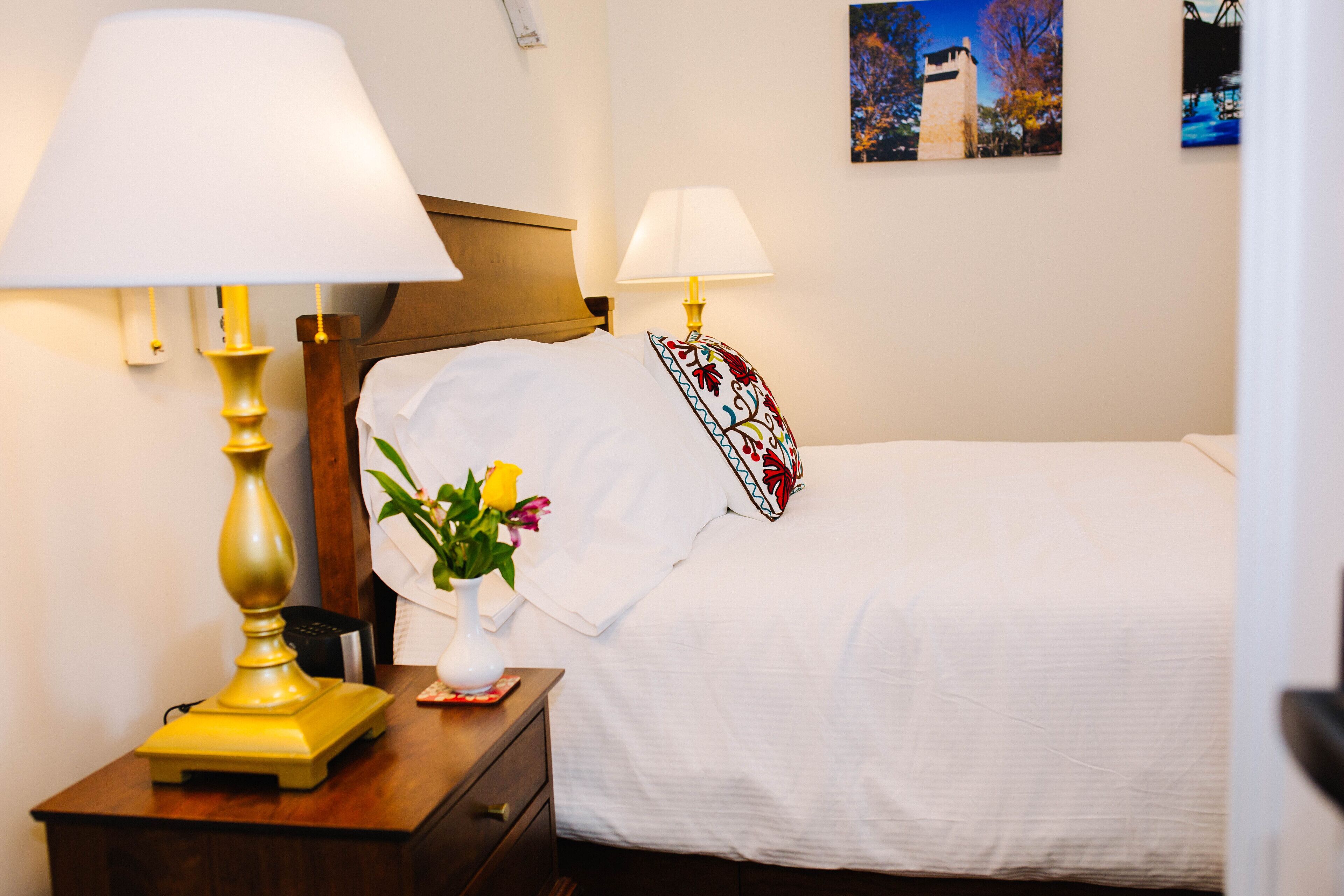 Un llit ben fet amb roba de llit blanca, un capçal de fusta i una almoinada decorativa. Hi ha una taula de nit amb una làmpada i un ram de flors.